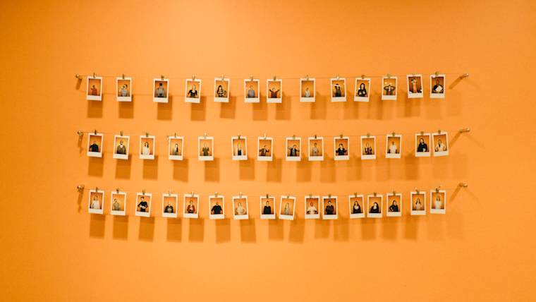 Rows of instant photos hung on strings against an orange wall, creating an organized display of portrait photographs