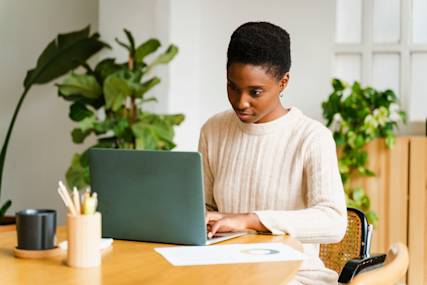 Person in cream sweater working on laptop at bright home office desk with houseplants and modern decor in background