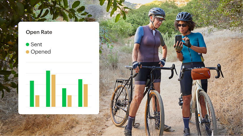 Two cyclists in athletic gear pause on a dirt trail to check their phones, with bikes and a scenic hillside backdrop